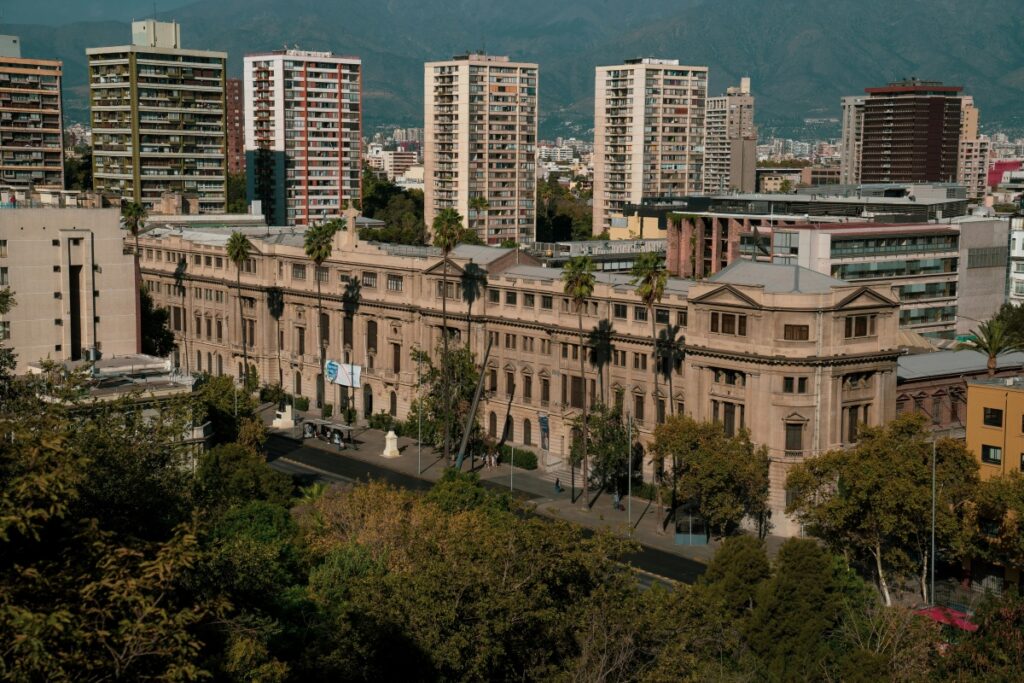 Esta imagen presenta una vista de una parte de la ciudad de Chile, donde un edificio histórico de estilo clásico se destaca entre modernos edificios altos. Rodeado de árboles, el edificio crea un bonito contraste con el paisaje urbano y la cordillera de montañas en el fondo. Para aquellos interesados en el marketing digital en Chile, las estrategias de "Facebook Ads en Chile Estrategias para Vender Más" pueden ayudar a promocionar esta clase de atractivos locales, atraer visitantes y aumentar las ventas de servicios relacionados con el turismo y la cultura.