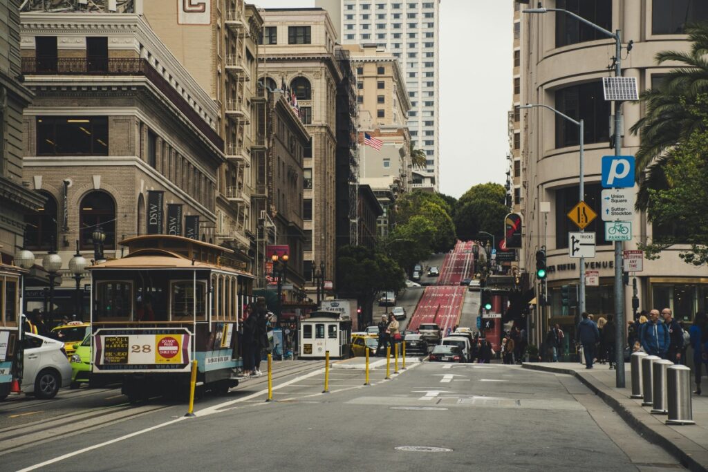Esta imagen muestra una calle en una ciudad con edificios históricos y modernos, un tranvía en movimiento y mucha gente. Además, se ve la pendiente de una colina. Al igual que esta calle atrae a muchas personas por su actividad y diversidad, en Instagram en Chile, para ganar más seguidores, debes crear contenido atractivo que muestre la esencia de la ciudad y de Chile, como sus monumentos, su cultura y su vida cotidiana. Utiliza HASHTAGS locales y populares, interactúa con la comunidad y publica regularmente para llamar la atención de nuevos seguidores.