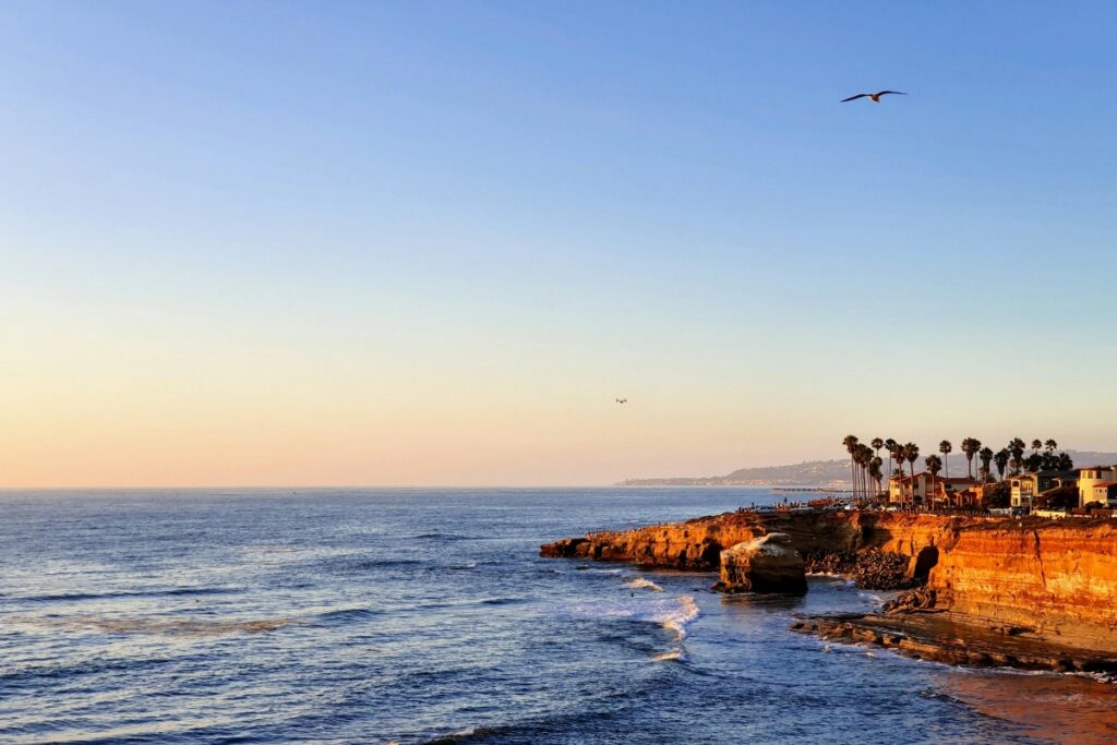 Esta es una hermosa vista del océano con olas que se rompen contra acantilados. En la parte superior, un ave vuela libremente. Cerca de los acantilados, hay edificios con palmeras. Esta imagen está relacionada con TikTok Chile Claves para Hacer Videos Virales. Aquí, la belleza natural puede inspirar a los creadores de contenido en TikTok Chile a crear videos atractivos y quizás hacer que sus videos se vuelvan virales, mostrando la maravilla de paisajes como este.
