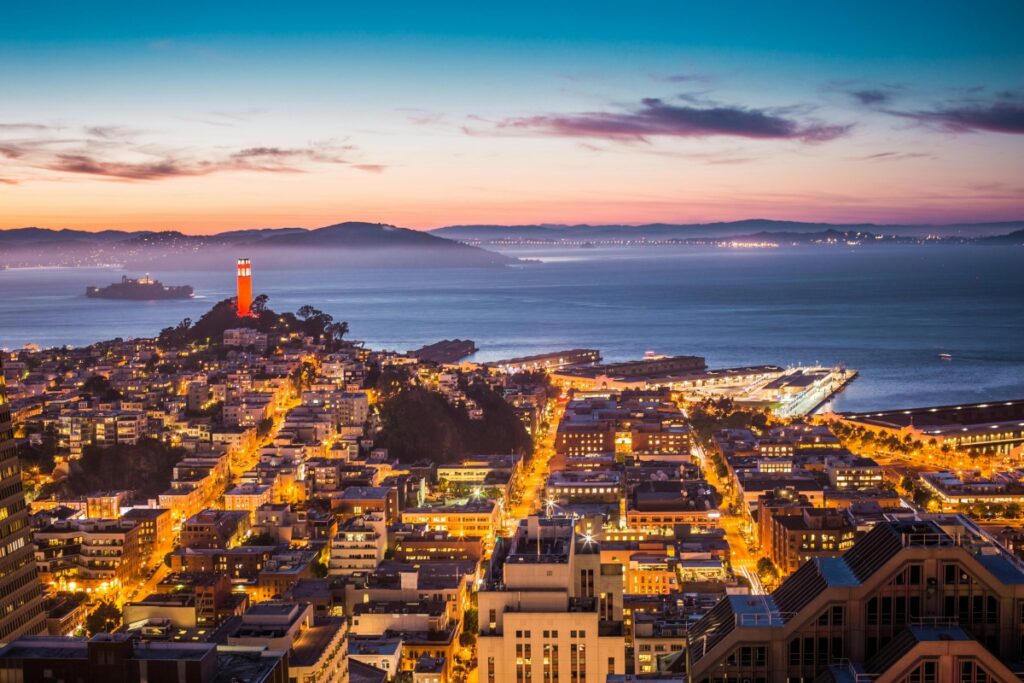 Esta imagen presenta una vista impresionante de una ciudad costera al atardecer. La ciudad está iluminada, y se pueden ver edificios, calles con luces y un faro rojo que destaca. Al fondo, el mar y una isla. Para aquellos en Chile que quieren ganar dinero a través de streaming en Twitch, mostrando paisajes como este y hablando sobre viajes y la belleza de ciudades costeras, siguiendo las estrategias de "Twitch en Chile Cómo Ganar Dinero con Streaming", pueden atraer a una audiencia y obtener ingresos a través de sus transmisiones.
